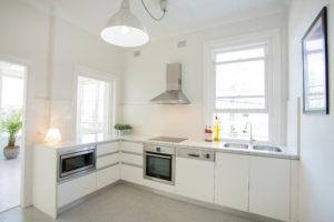 Design ideas for a small contemporary l-shaped separate kitchen in Sydney with a double-bowl sink, flat-panel cabinets, white cabinets, solid surface benchtops, white splashback, porcelain splashback, stainless steel appliances, laminate floors, no island and grey floor.